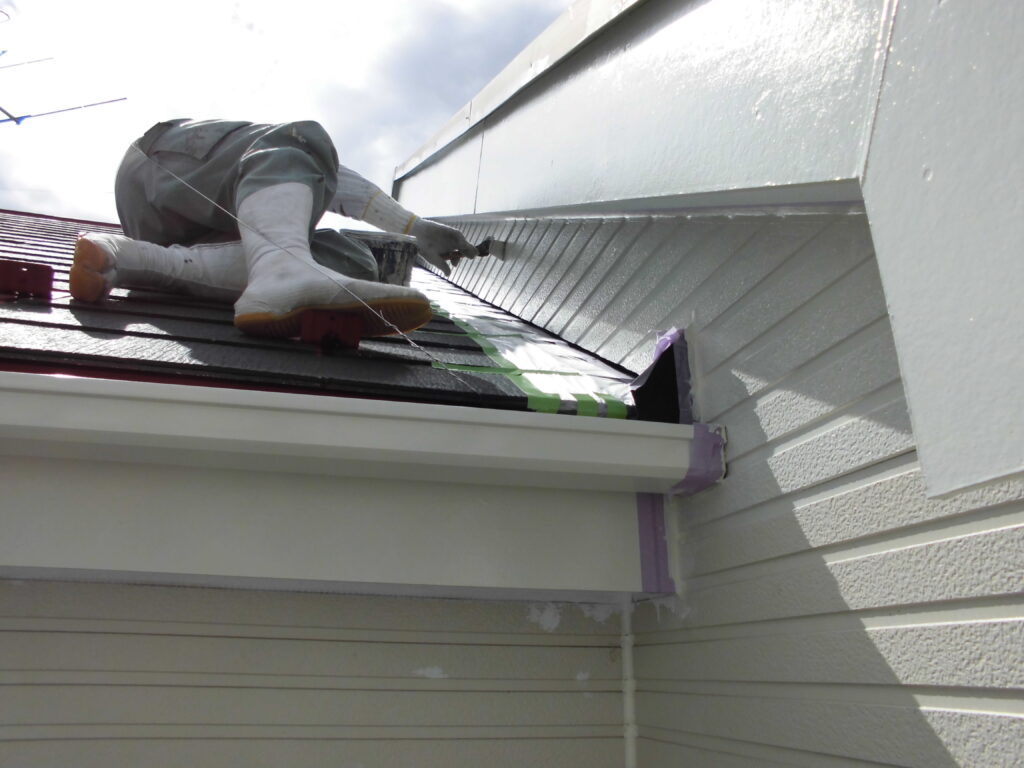 Construction worker in white boots kneels on a sloped black metal roof, applying sealant to the edge near the gutter of a house