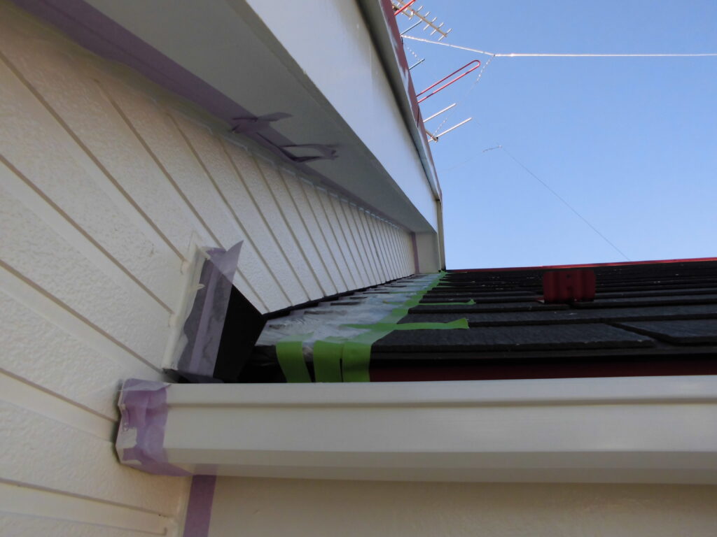 Close-up of a house eave under repair with masking tape along the trim and roof edge, blue sky above, and a piece of guttering visible.
