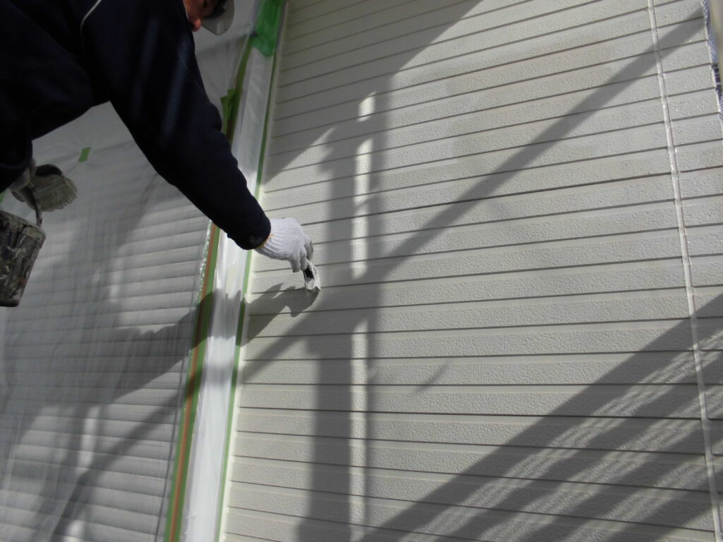 A painter wearing a glove applies white paint to exterior siding along a taped edge in progress on a house.