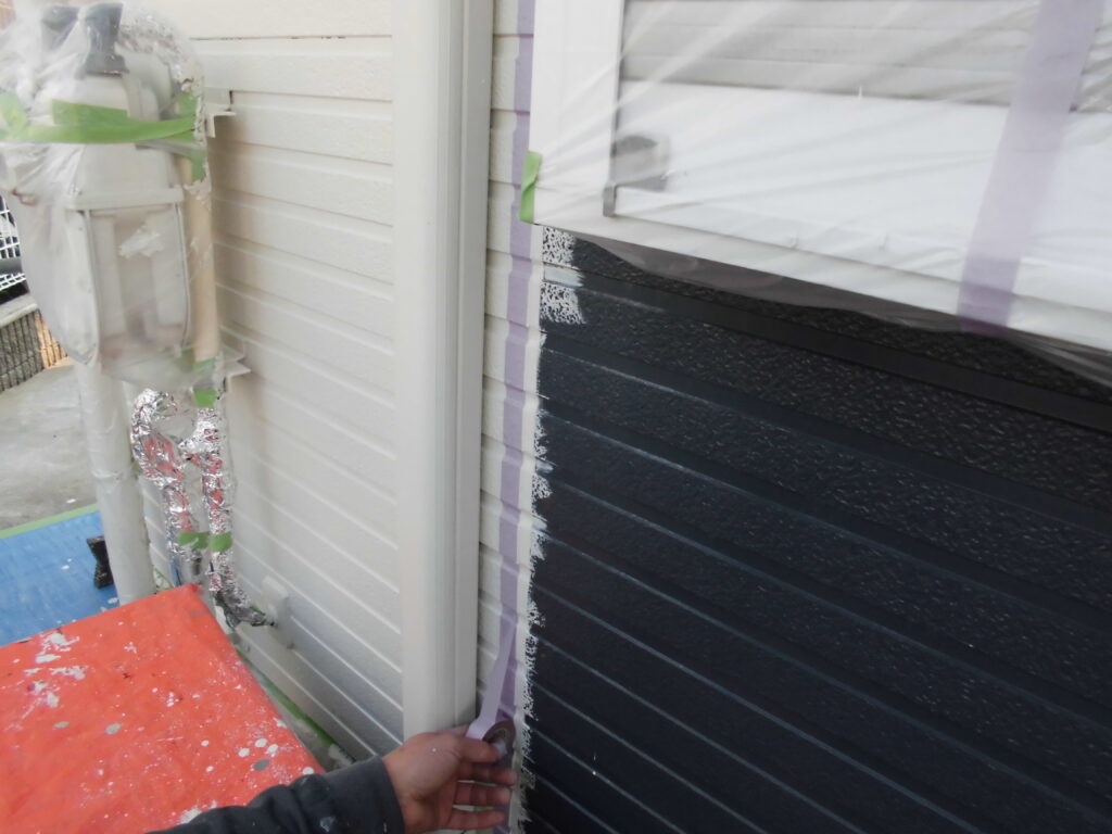 Worker applying painter’s tape along the seam where light siding meets dark blue exterior during exterior painting, with a window protected nearby.
