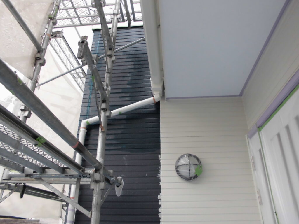 Metal scaffolding along a house exterior, with white and dark siding, a round wall vent, and a white drain pipe visible