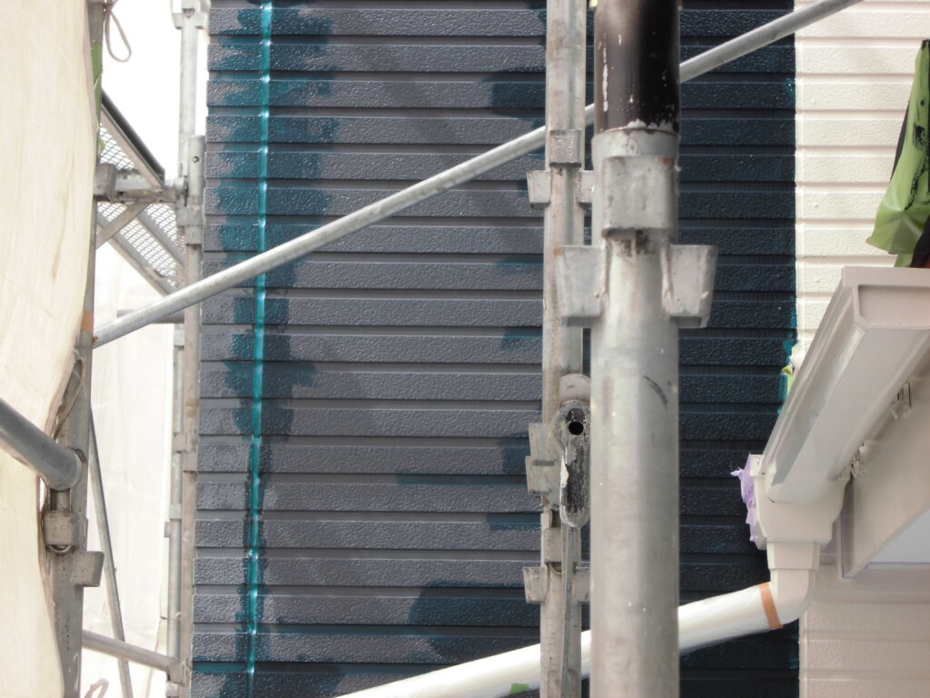 Construction scaffolding in front of a dark blue corrugated wall with metal poles and clamps, a vertical teal line, and a white gutter downspout on the right.
