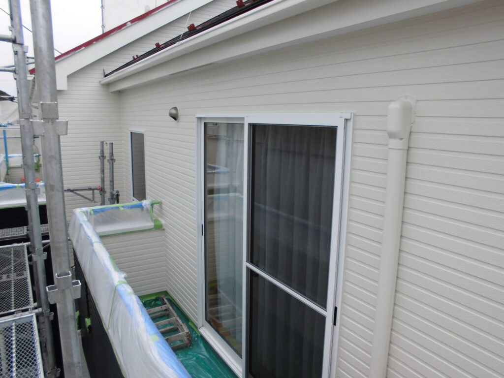 Construction scene: beige exterior siding, sliding glass door, and scaffolded balcony with protective plastic cover on the railing