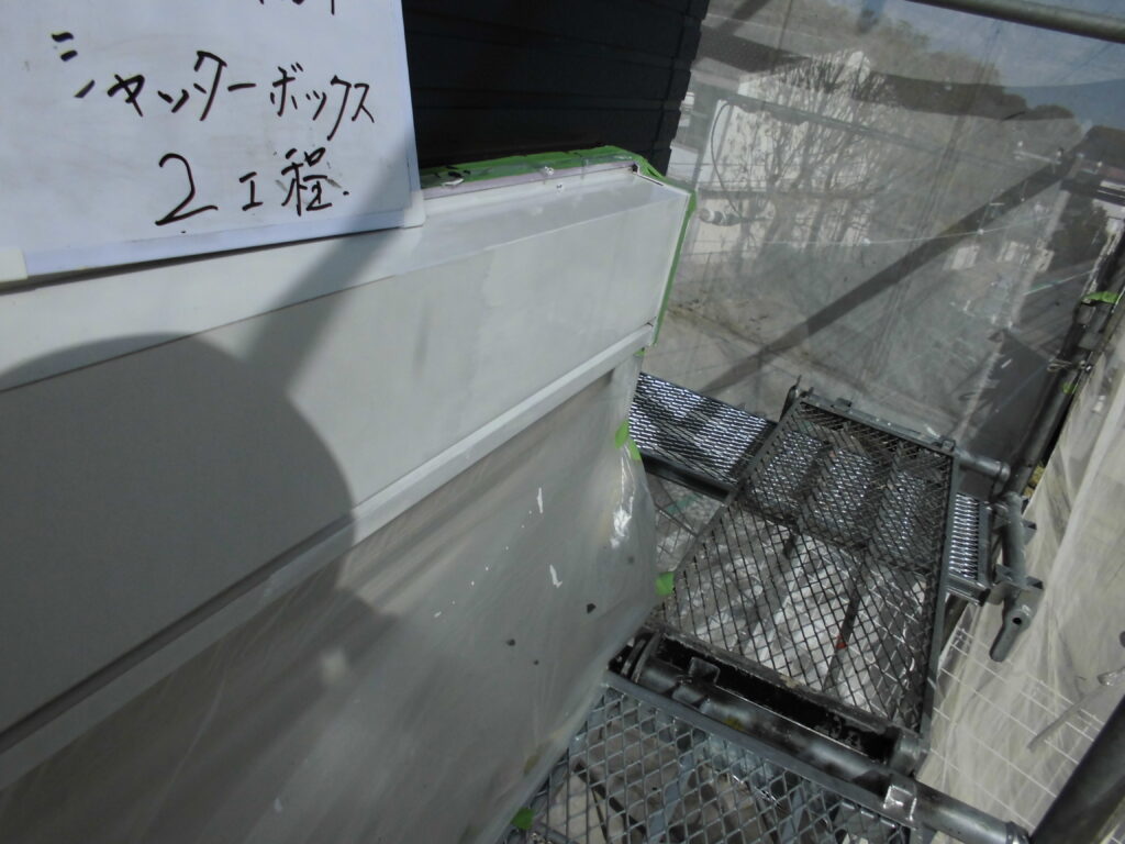 White sign with Japanese writing attached to a gray metal surface, at a construction scaffold with metal grating platform visible in the background from a side angle.