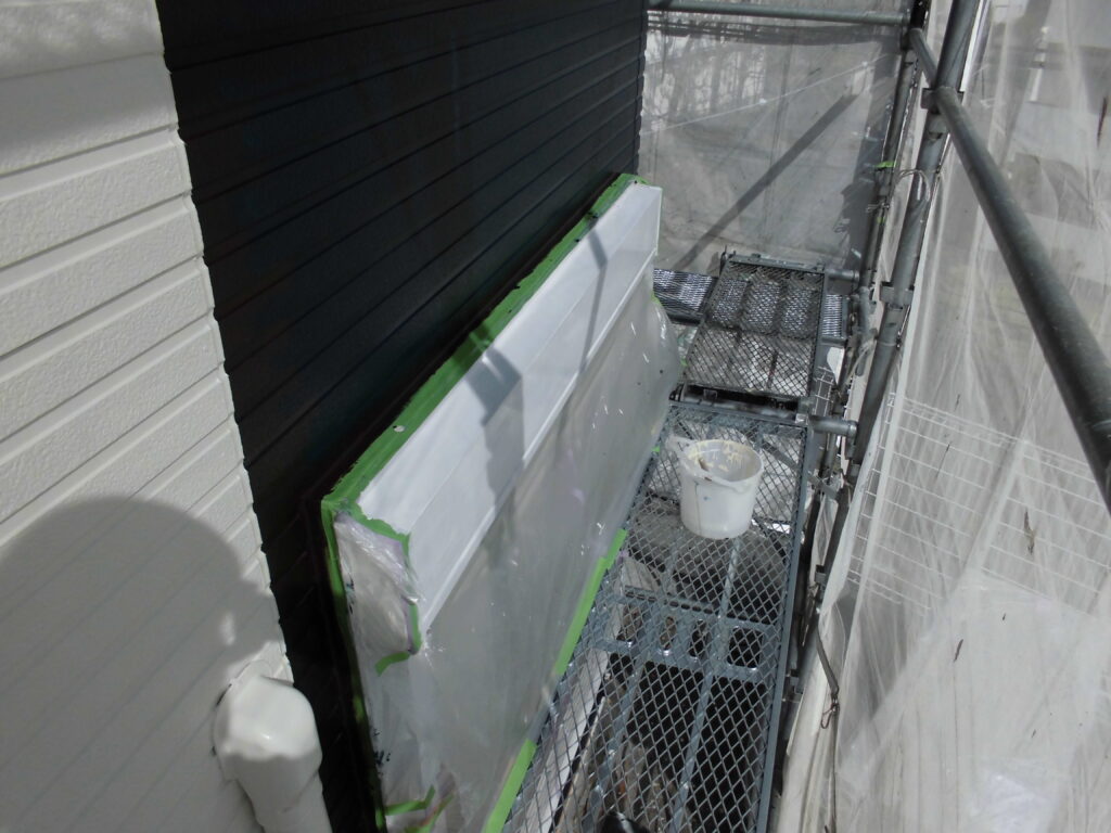 Narrow scaffold platform beside a white exterior wall; a large panel masked with green tape is prepped for painting, with a white bucket on the grate.