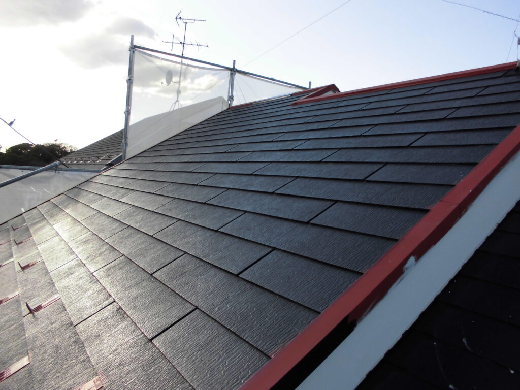 Sloped roof with dark slate shingles and red edge trim; scaffolding with a glass panel visible in the background.