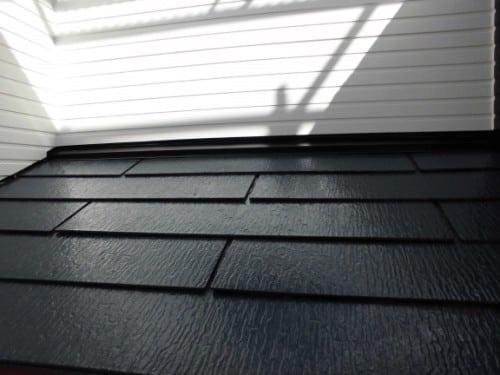 Close-up of dark asphalt roof shingles along a building edge, with a white siding wall and strong diagonal shadows overhead.