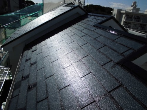 Sloped rooftop covered with dark shingles, sunlight reflecting off a textured surface near scaffolding and adjacent buildings.