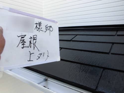 Close-up of a hand holding a white sign with handwritten Japanese characters, in front of a dark shingled roof.