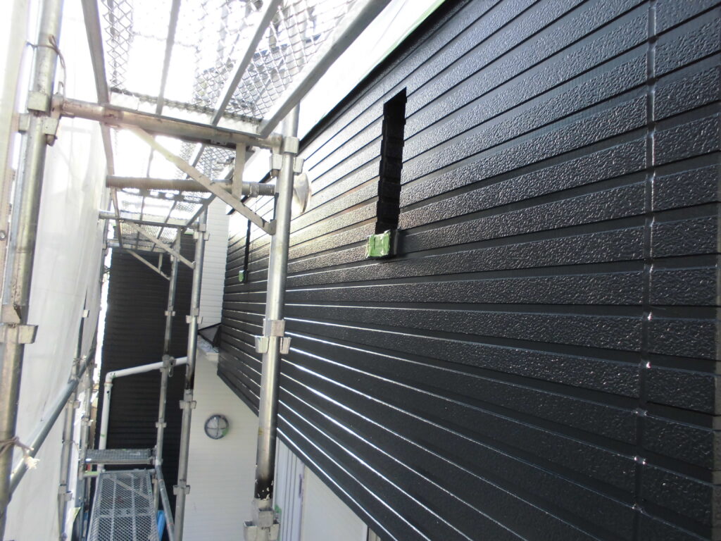 Scaffolding runs along a dark textured exterior wall with a rectangular opening in the siding, viewed from a narrow alley between buildings.