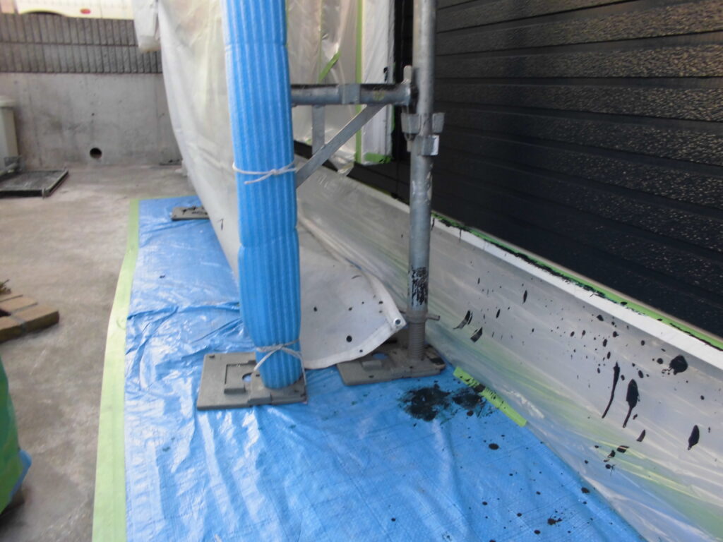 Scaffolding beside a building with a blue wrapped pole, blue tarp on the ground, and paint splatters along the white base trim.