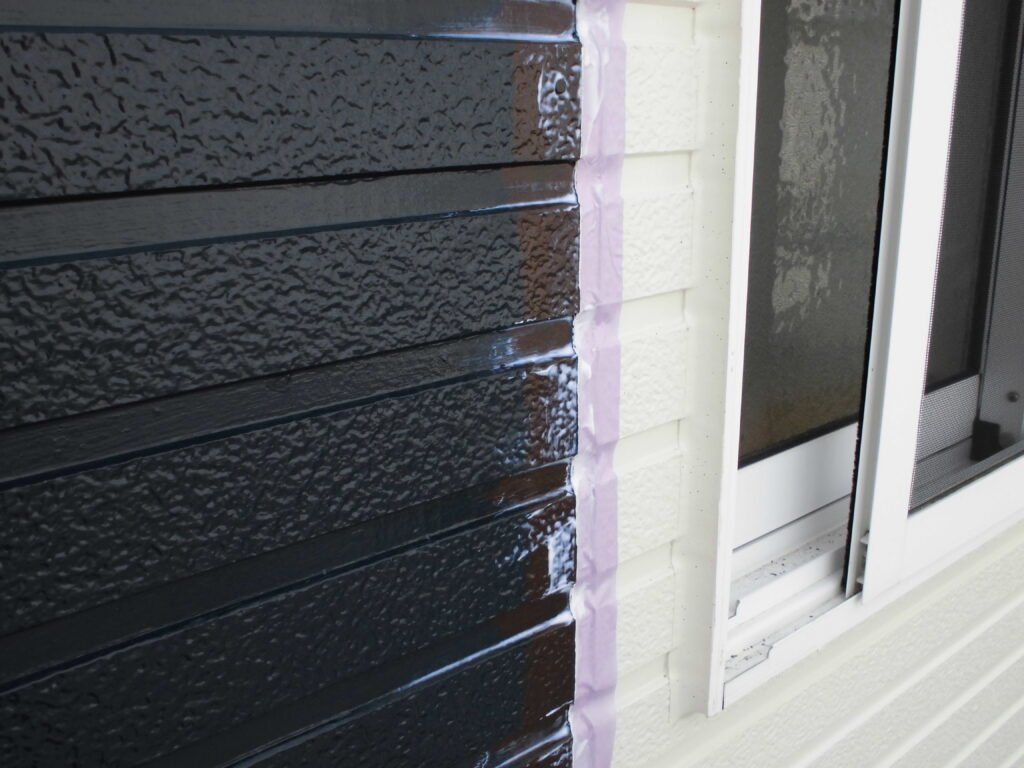 Close-up of dark brown textured siding being painted along a white house wall with pink masking tape beside a window.