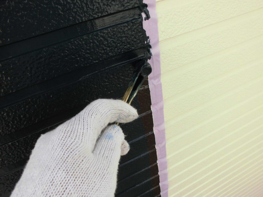 Gloved hand painting black trim with a small brush along masked edge of a yellow wall outside.