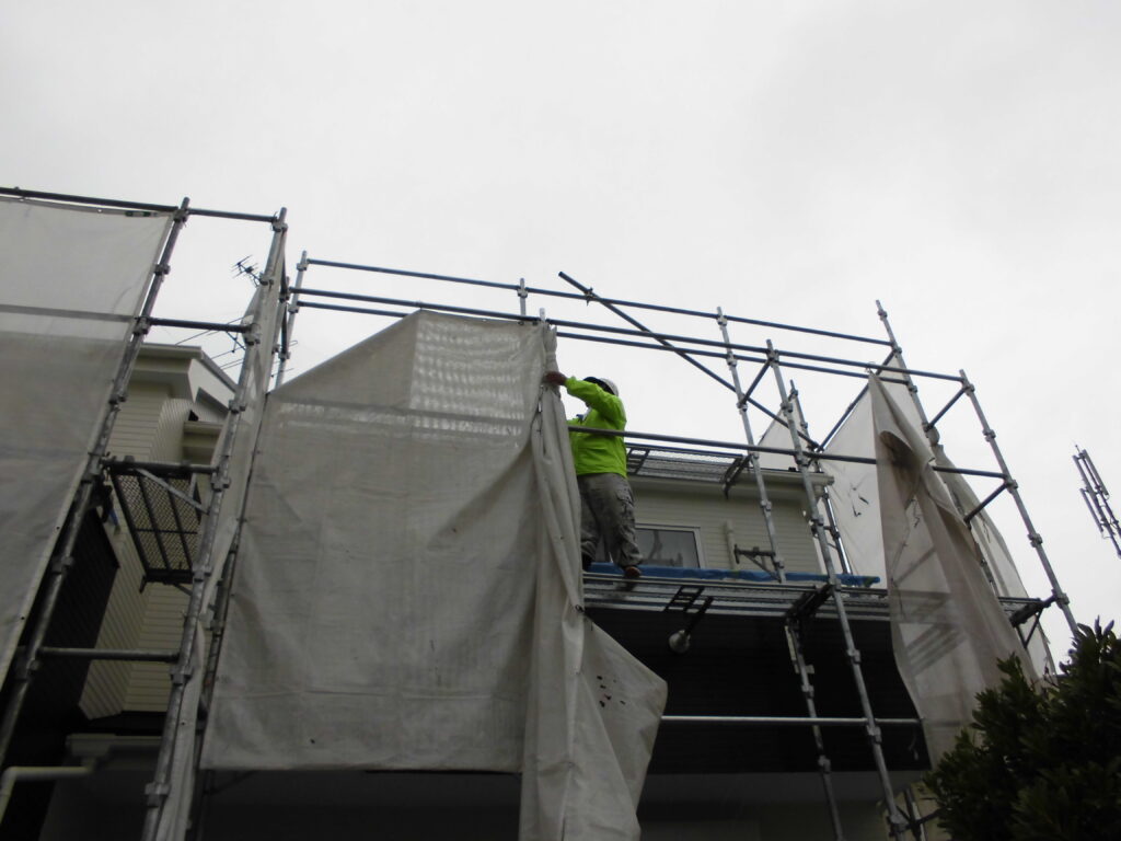 Construction worker in a bright green shirt stands on scaffolding around a two‑story house, handling white protective tarps.