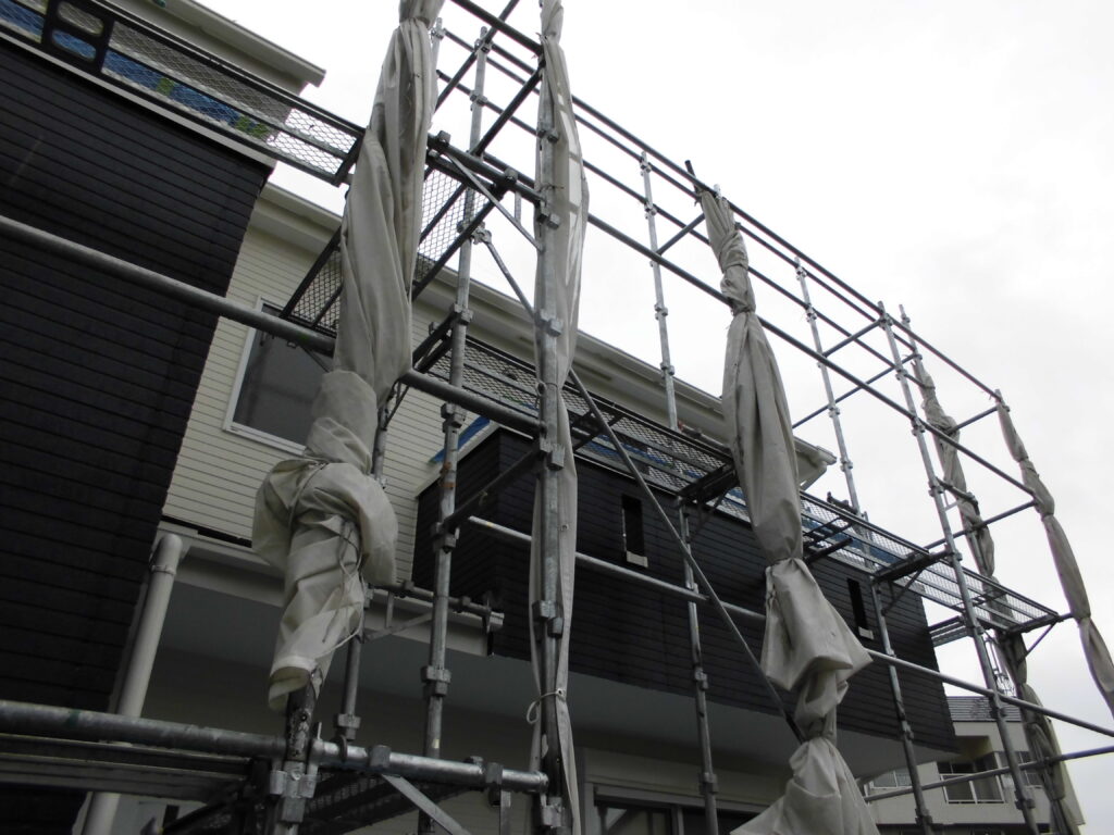 Construction scaffolding surrounds a modern two-story house under renovation, with wrapped poles and safety nets visible.