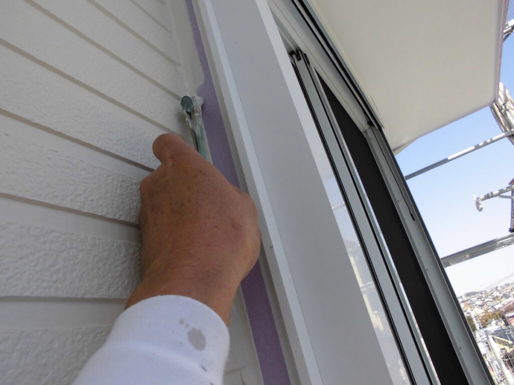 Close-up of a hand applying purple sealant along the seam beside a window on white siding exterior wall. (informative)