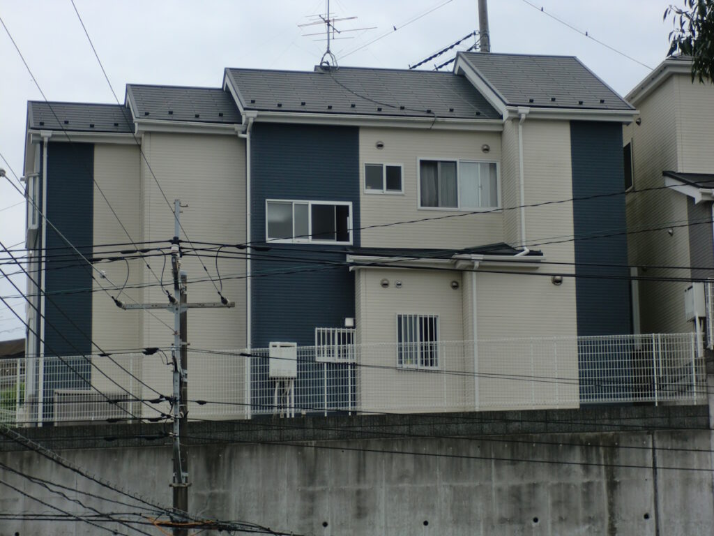 Three-story beige and dark-blue apartment building with multiple windows, behind a fence and tangled overhead power lines.