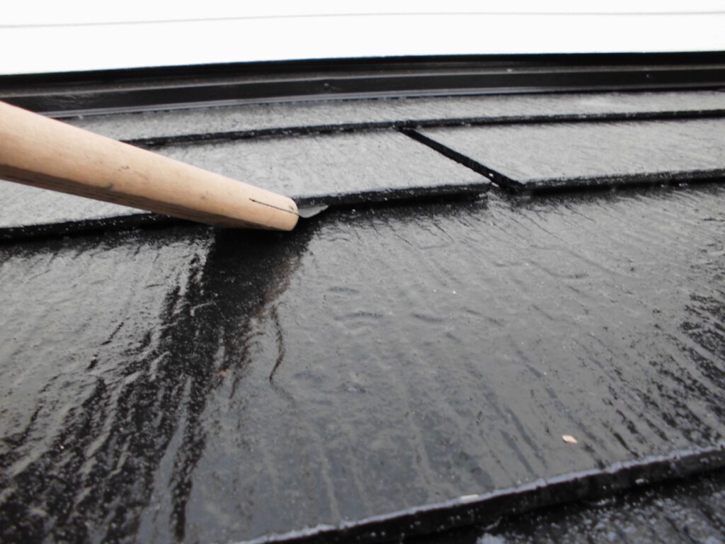 Close-up of a wooden tool pressing on wet black roof shingles beneath a white gutter, showing construction work in progress.