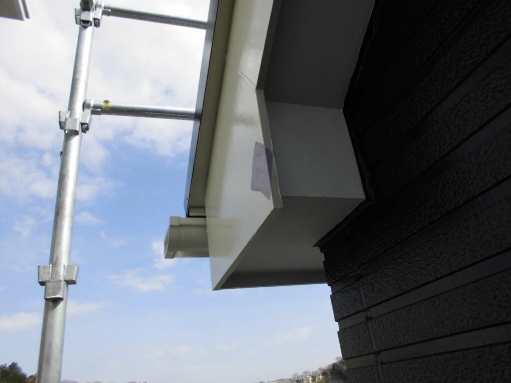 Metal scaffolding pole beside a white house cornice, with dark textured siding on the right against a blue sky.