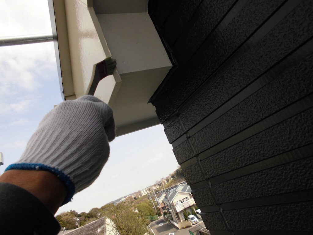 Gloved hand holding a small brush painting the underside of a building's eaves, with a street scene below.