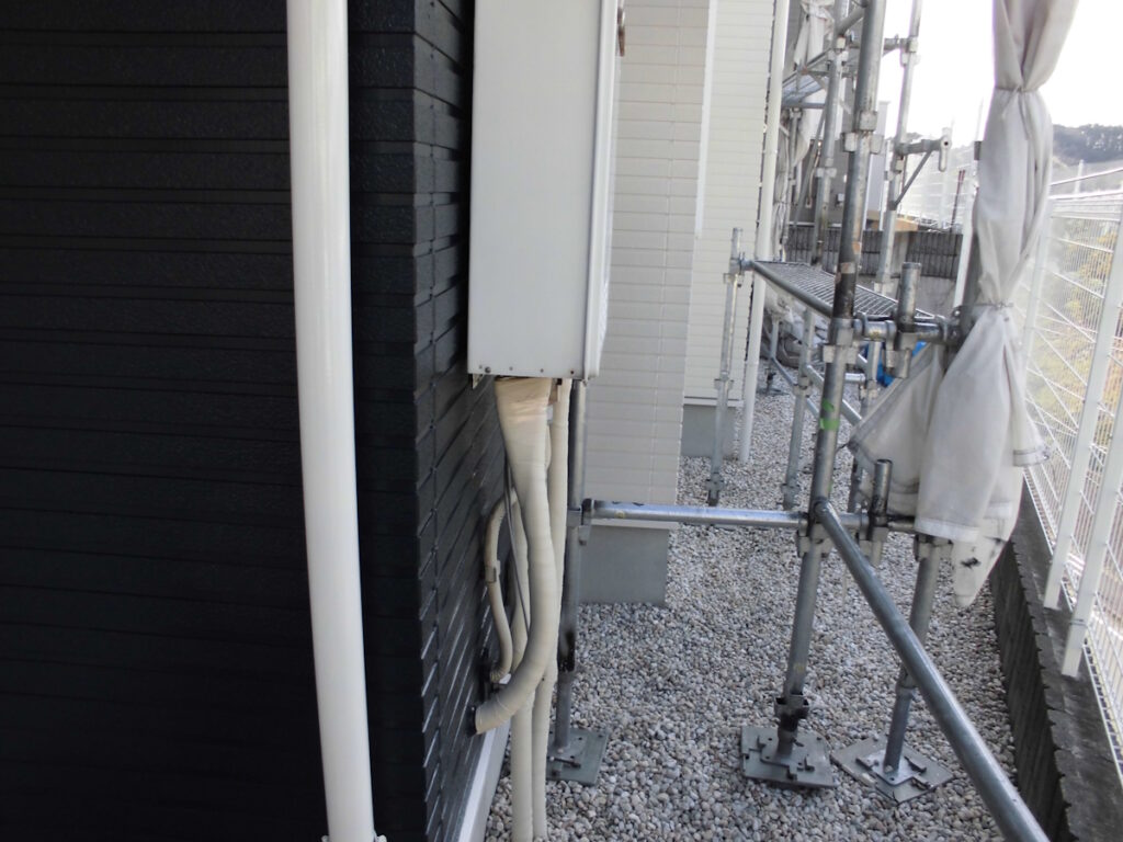 Exterior construction scaffolding runs along the side of a building, with a white utility box and bundled pipes attached to a dark wall on a gravel surface.