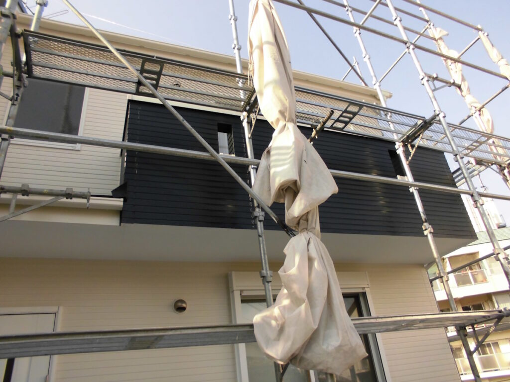 Scaffolding surrounds a modern two-story house with dark siding and beige fabric wraps hanging from the framework.