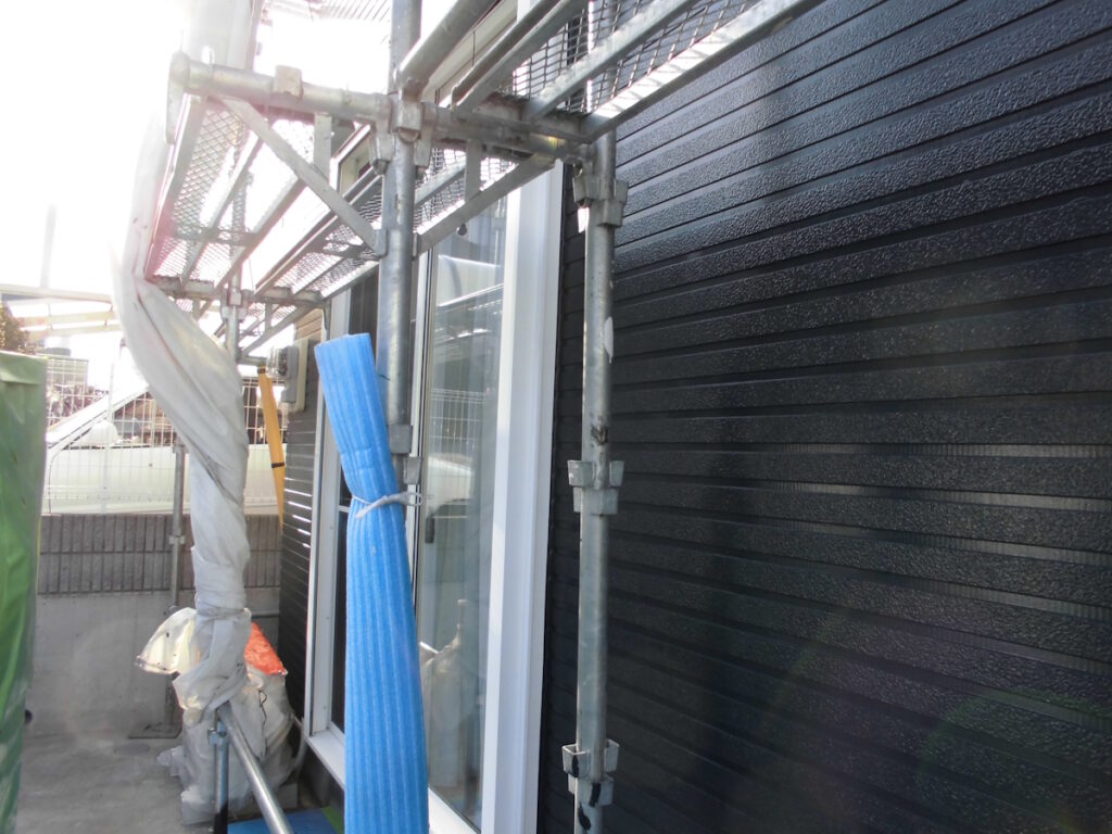 Scaffolding along the side of a building with black textured siding; a blue protective padding/pipe is visible near a window.