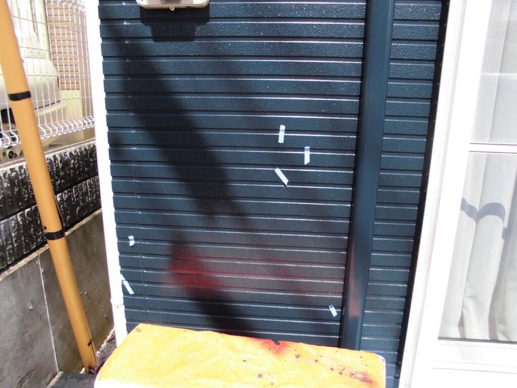 Blue corrugated metal exterior with several white tape strips, yellow post on the left, and an orange bench in the foreground near a window with curtains on the right.