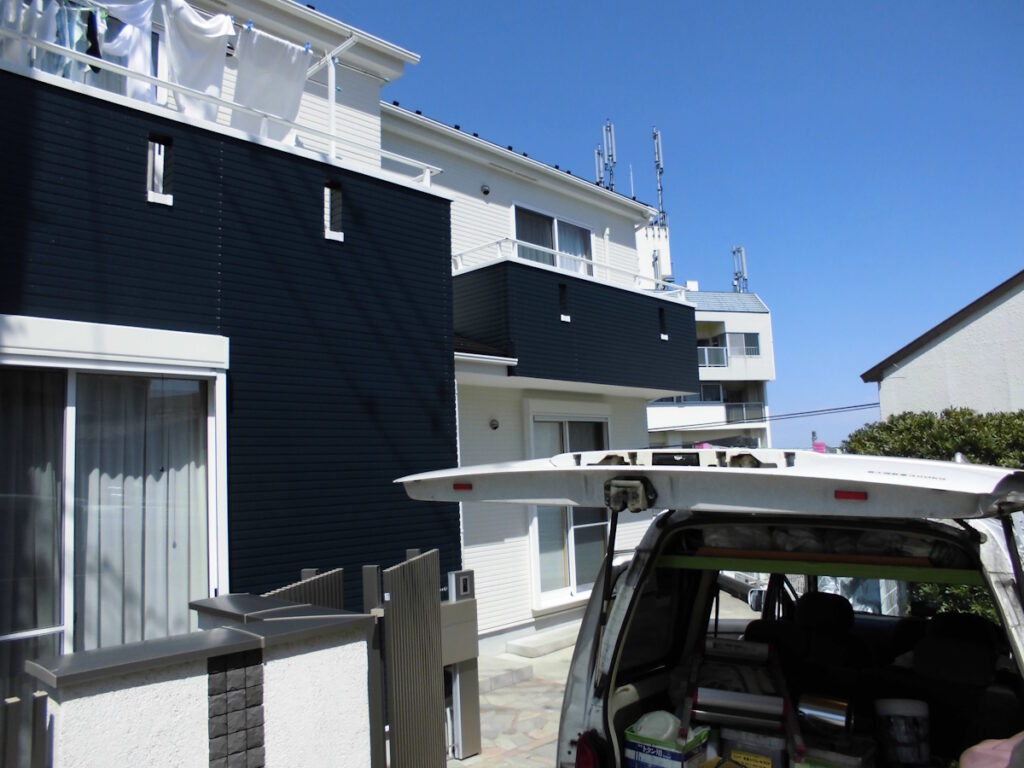 Apartment building with dark blue siding and laundry hanging on the top balcony; a van with its rear hatch open in front.
