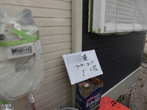 Exterior wall of a building under renovation: plastic-wrapped utilities/meter and a handwritten Japanese sign propped on a box near the ground.