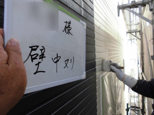 Construction worker wearing a glove paints a dark exterior wall with a roller; a whiteboard with Japanese writing is held up nearby.