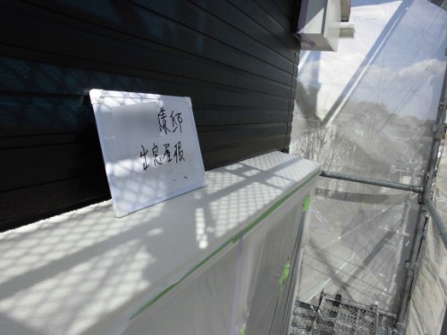 Whiteboard with handwritten Japanese text rests on a white ledge against a dark exterior wall, with scaffolding and clear plastic sheeting in a sunlit construction area.