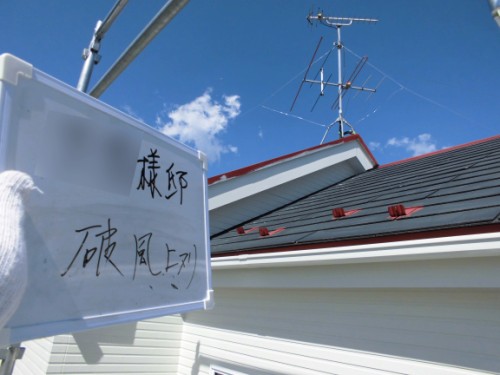 White sign with handwritten Japanese text attached to a metal frame on a rooftop under a blue sky and antennae in the background.