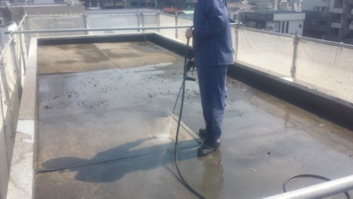 20140317 _104217 | 外壁塗装は神奈川県横浜市の塗装職人 Person in a blue work uniform uses a pressure washer to clean a flat rooftop with puddles on the surface.