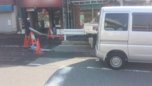 20140423-083544 | 外壁塗装は神奈川県横浜市の塗装職人 Construction scene on a city street: orange cones block the sidewalk as a long metal duct is laid across the pavement, a worker nearby, and a white van parked on the road.
