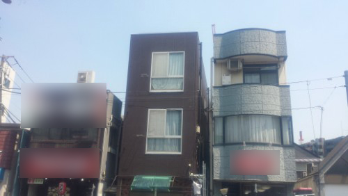 20140423-114052 | 外壁塗装は神奈川県横浜市の塗装職人 Three adjacent urban buildings on a sunny street: a narrow dark brown three-story house in the center, with a blue rounded-corner building to the right and blurred storefronts to the left.