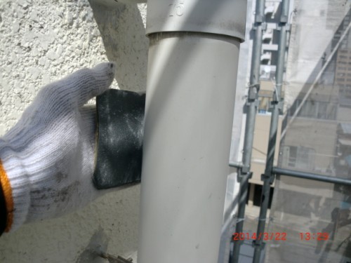 CIMG0376 | 外壁塗装は神奈川県横浜市の塗装職人 Gloved hand applying a dark patch to a rough wall beside a gray vertical pipe at a construction site.