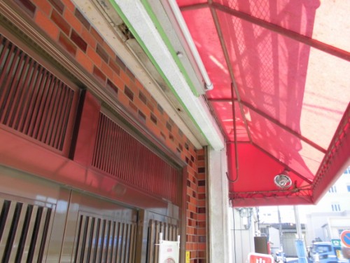 CIMG1386 | 外壁塗装は神奈川県横浜市の塗装職人 Storefront with metal security shutters, brick wall, and a red fabric awning over the entrance.