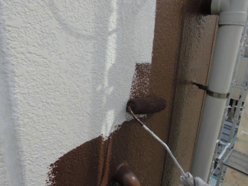 CIMG1390 | 外壁塗装は神奈川県横浜市の塗装職人 Close-up of a paint roller applying brown paint over a white textured exterior wall beside a vertical drain pipe during a repaint.