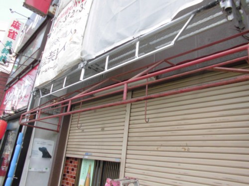 CIMG1399 | 外壁塗装は神奈川県横浜市の塗装職人 Closed beige storefront shutters under scaffolding and a fabric awning on a busy street.