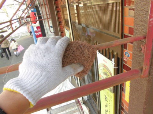 CIMG1422 | 外壁塗装は神奈川県横浜市の塗装職人 Gloved hand holding a brown scrub pad scrubbing a red metal railing on scaffolding outside a storefront.