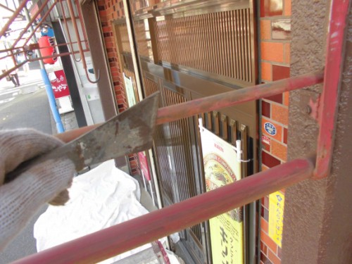 CIMG1423 | 外壁塗装は神奈川県横浜市の塗装職人 Gloved hand holding a scraper against a red metal railing outside a brick storefront, mid-scraping motion.