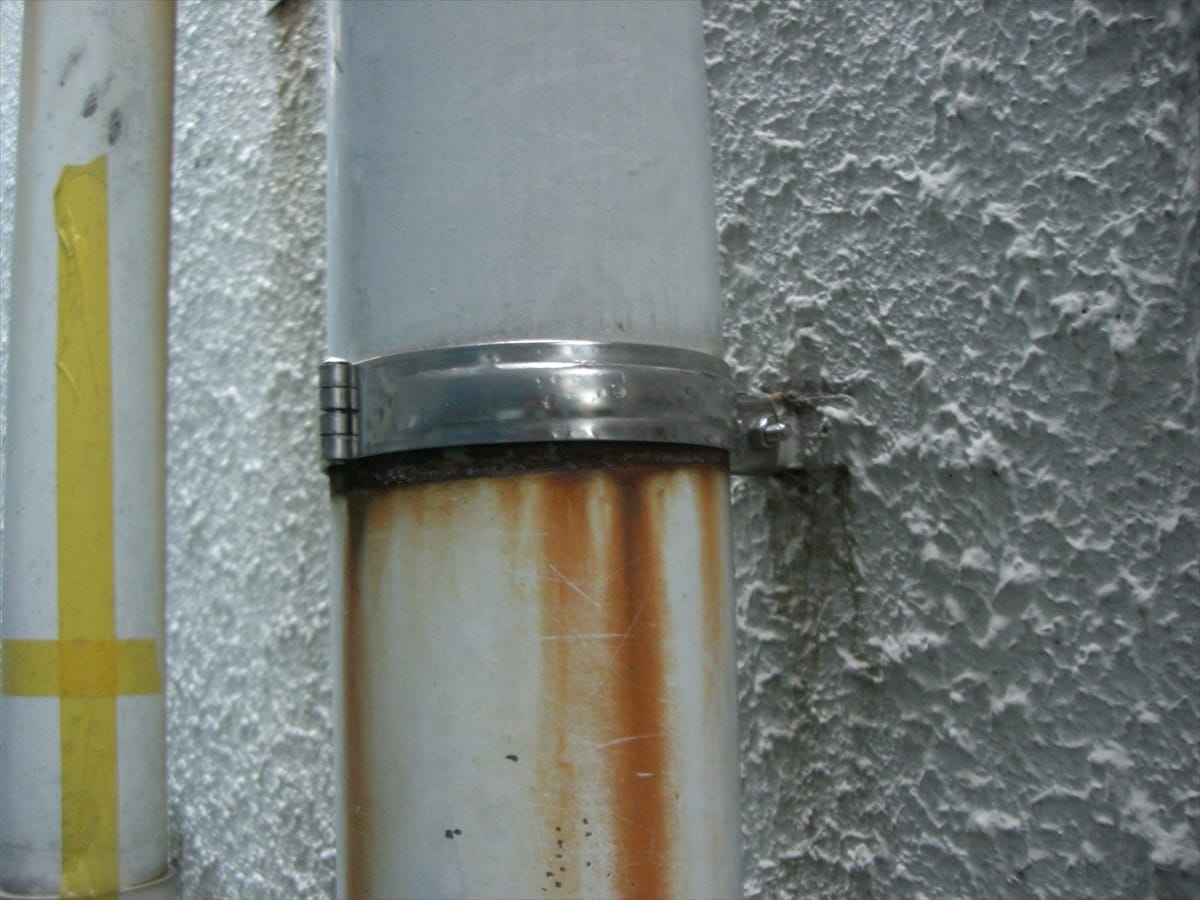 CIMG1582stainless | 外壁塗装は神奈川県横浜市の塗装職人 Close-up of a rust-stained vertical pipe with a metal clamp mounted to a textured gray wall.