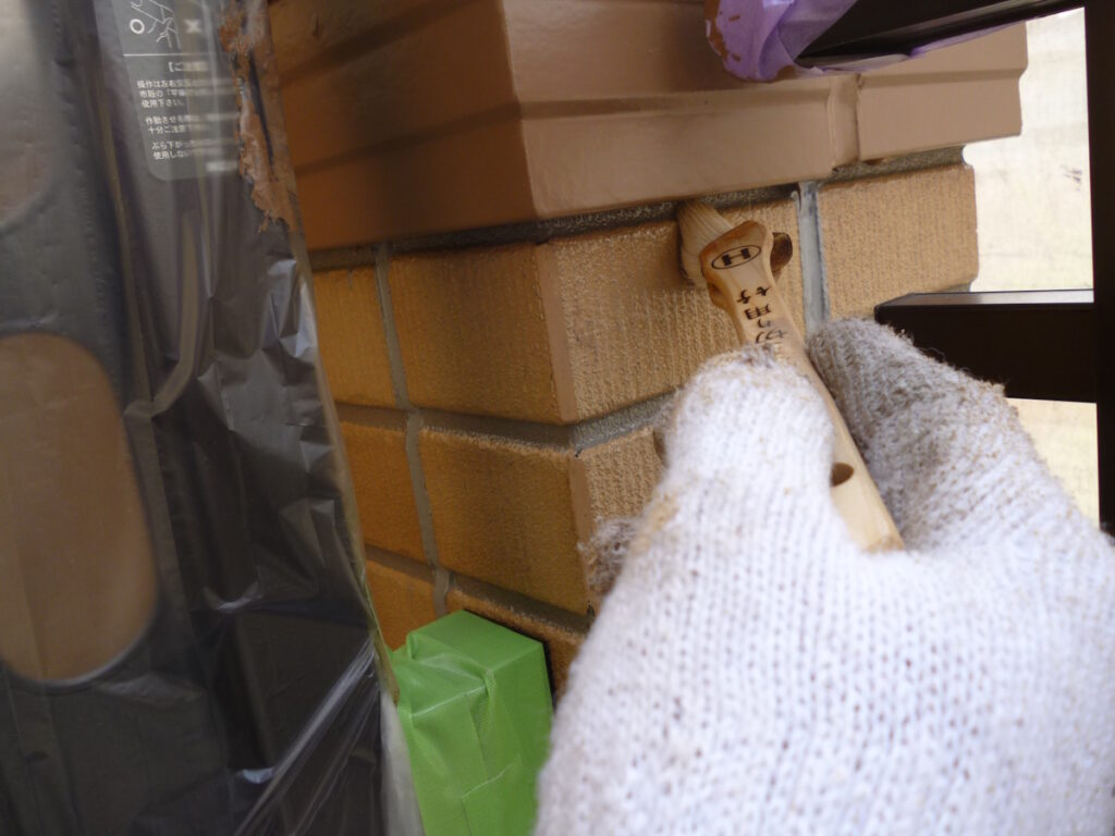 Gloved hand holding a wooden tool with writing, near a brick wall and stacked boxes in a cluttered area
