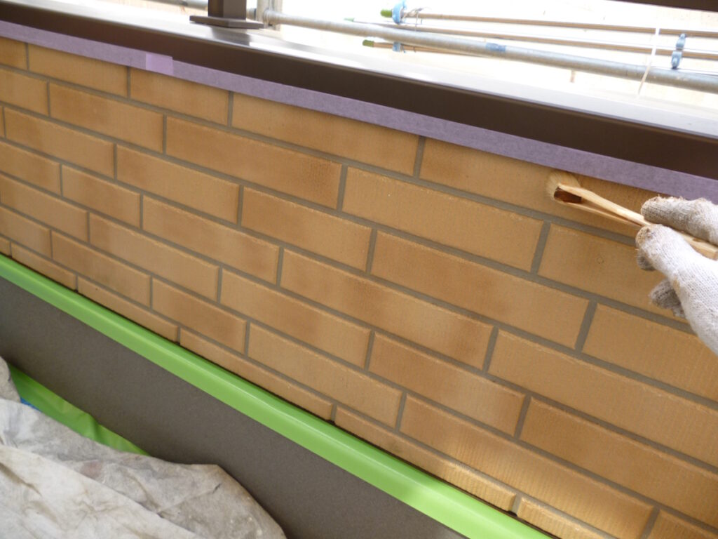 Construction worker wearing gloves uses a wooden shim on a brick exterior wall with green masking tape along the base.