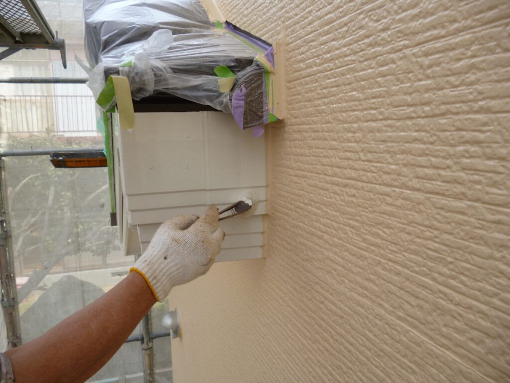 Worker in a glove paints a beige exterior vent cover on a textured wall, with masking tape and plastic sheeting nearby.
