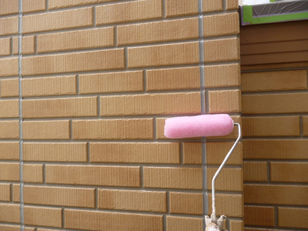 Pink paint roller pressed against a beige brick wall, mid-painting a section of the exterior surface.
