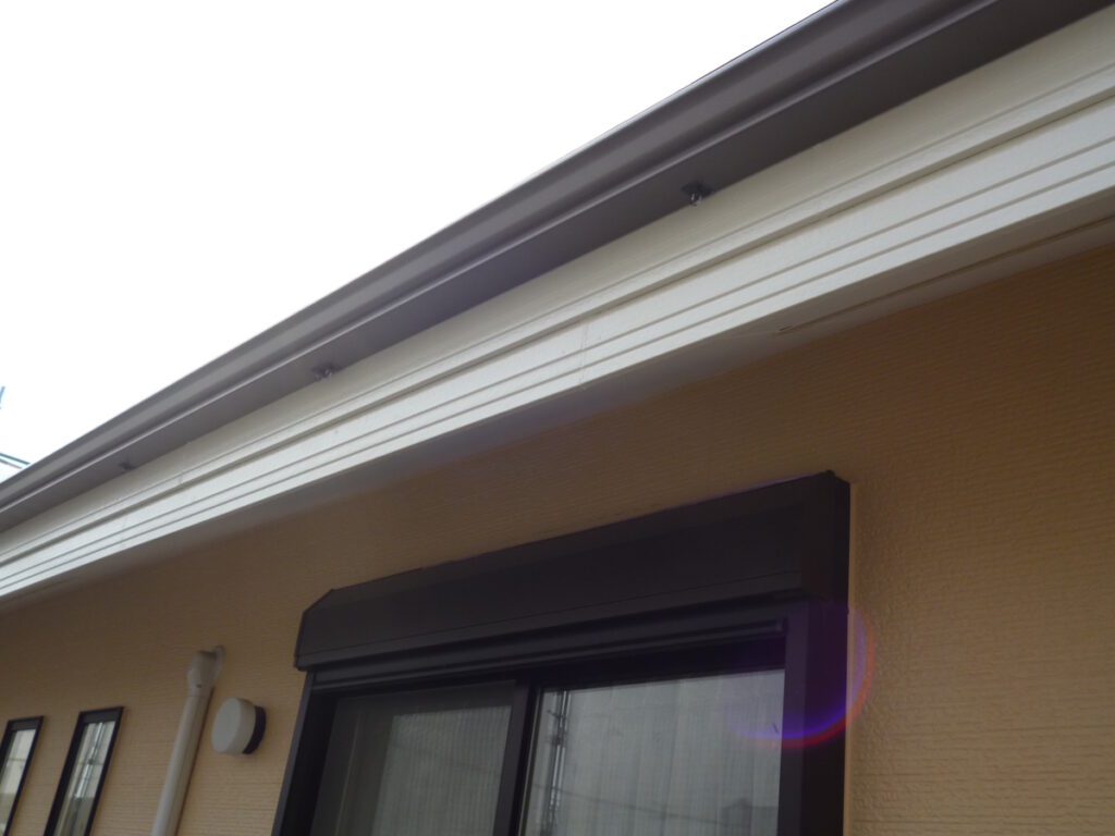 Beige house wall with a black-framed window and a closed exterior rolling shutter; white gutter along the top and a round wall light nearby.