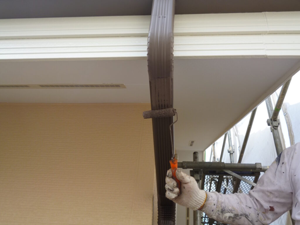 Painter rolling brown paint onto a vertical downspout on a building with scaffolding nearby.
