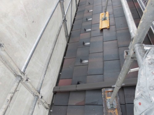 Sloped dark slate roof under construction, surrounded by scaffolding; a yellow tool block and a brown pipe lie across the tiles.